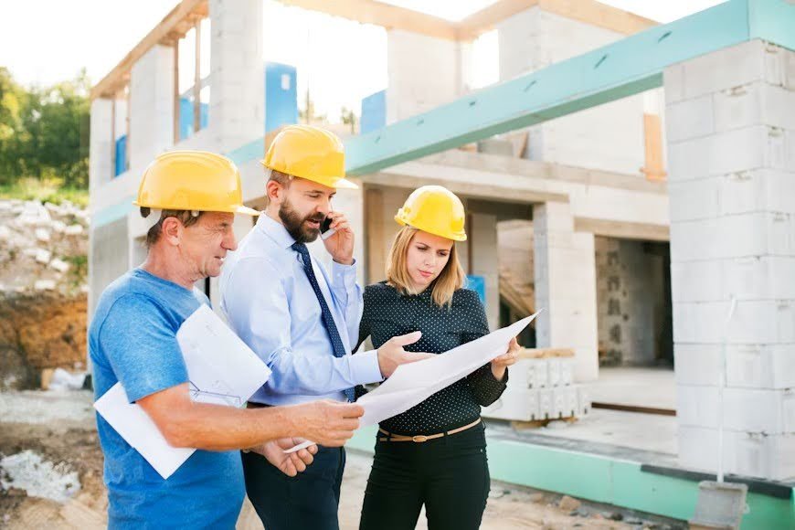A civil engineer and two workers review plans and blueprints while discussing issues at a construction site.