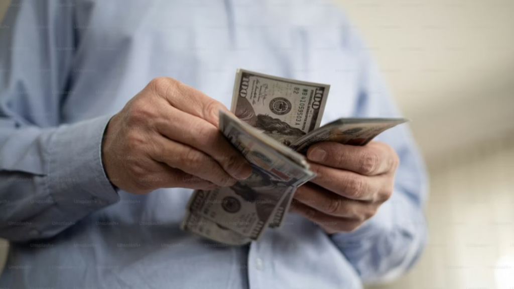 An adult man in a blue long-sleeve shirt is counting $100 bills at home.